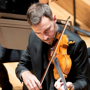 Simon Gilks is a white man with short brown hair. He is playing the violin during a concert and wearing a black suit jacket and black shirt.