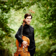 A white man with brown hair wearing a black shirt and black trousers, holding a cello and standing in a forest.