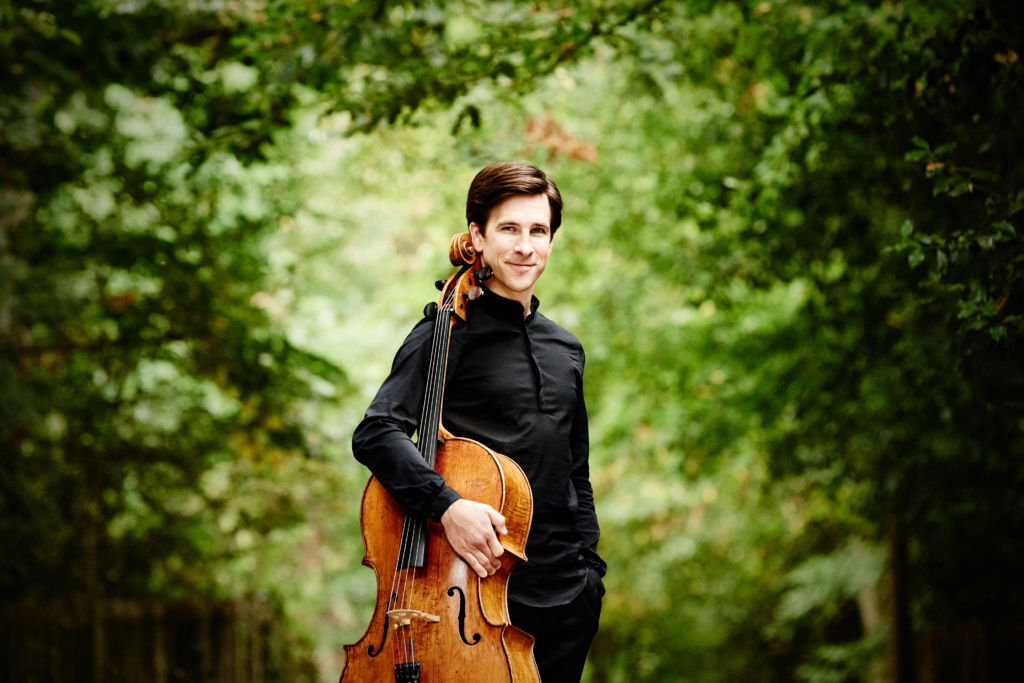 A white man with brown hair wearing a black shirt and black trousers, holding a cello and standing in a forest.