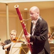 Ben Hudson playing the bassoon during a concert. Ben is a white man with a bald head and he is wearing a black tie outfit with a white shirt.