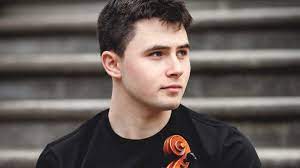 A white teenage boy with brown hair, wearing a black tshirt and holding a viola. 
